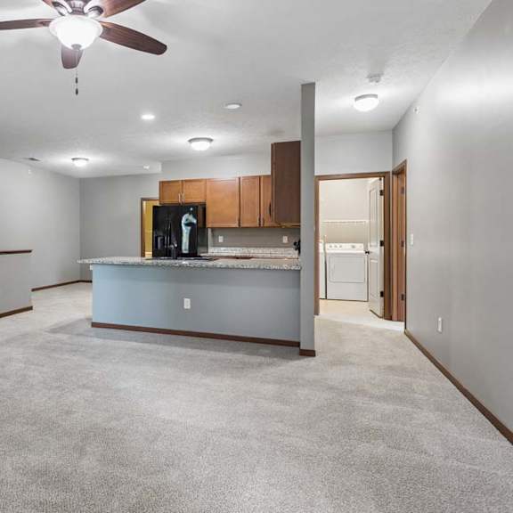 Thumbnail 7 of 16 Open-concept apartment living area with carpet, ceiling fan, breakfast bar, wood cabinetry, black refrigerator, and laundry room access.