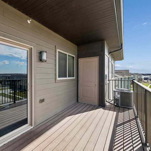 Thumbnail 44 of 44 Covered apartment balcony with wood-style decking, a large sliding glass door, exterior lighting, and metal railings overlooking the surrounding community.