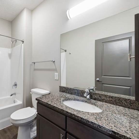 Thumbnail 42 of 44 Apartment bathroom with a granite-look single-sink vanity, dark cabinetry, a large mirror, and a tub-shower combination with a white surround and modern fixtures.