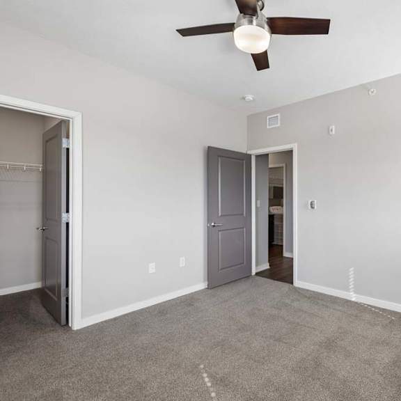 Thumbnail 39 of 44 Spacious apartment bedroom with carpeted flooring, a ceiling fan, a walk-in closet, and a window with blinds providing natural light.