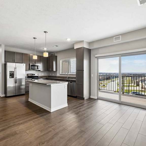 Thumbnail 24 of 44 Spacious open-concept apartment living area with wood-style flooring, a modern kitchen featuring a center island, pendant lighting, stainless steel appliances, and a large sliding glass door leading to a private balcony.