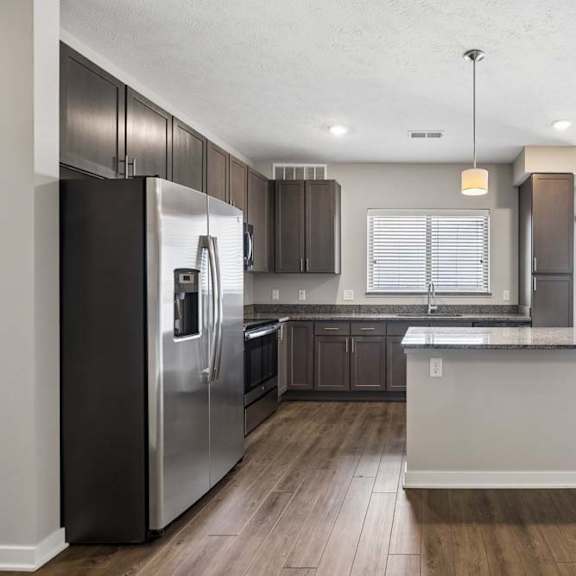 Thumbnail 25 of 44 Open-concept apartment kitchen with a large island, dark cabinetry, granite-look countertops, stainless steel appliances, pendant lighting, and a spacious living area with a sliding glass door leading to a private balcony.