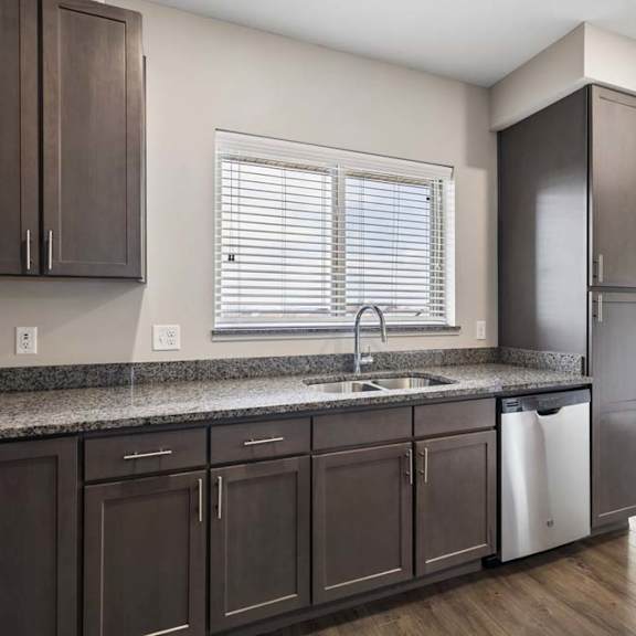 Thumbnail 28 of 44 Modern apartment kitchen with dark cabinetry, granite-look countertops, a stainless steel dishwasher, and a large window above the sink providing natural light.