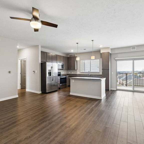 Thumbnail 26 of 44 Spacious open-concept apartment living room with wood-style flooring, a ceiling fan, and a modern kitchen featuring a breakfast bar, dark cabinetry, stainless steel appliances, and a large sliding glass door leading to a private balcony.