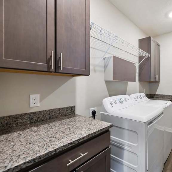 Thumbnail 43 of 44 Apartment laundry room with side-by-side washer and dryer, granite-look countertop, dark cabinetry, built-in wire shelving, and wood-style flooring.
