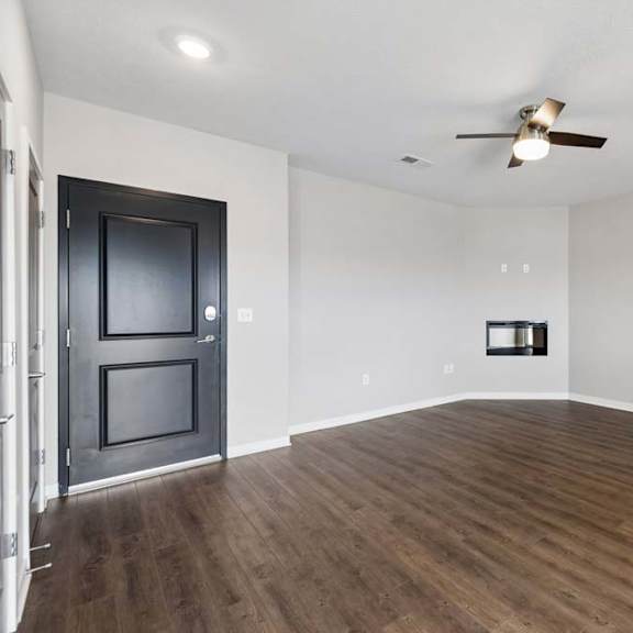 Thumbnail 31 of 44 Spacious apartment living room with wood-style flooring, a ceiling fan, a wall-mounted fireplace, and an open layout with hallway access and a dark entry door.