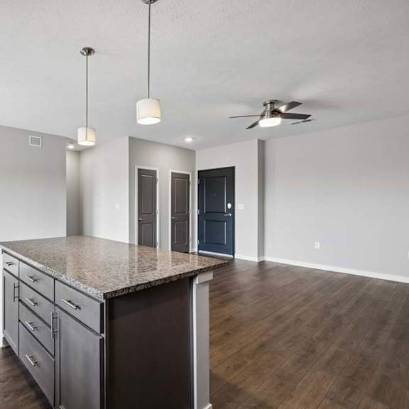 Thumbnail 27 of 44 Open-concept apartment kitchen with a large island, granite-look countertops, pendant lighting, and dark cabinetry, flowing into a spacious living area with wood-style flooring and a ceiling fan.