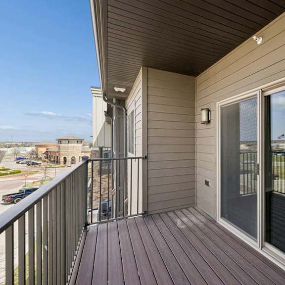 Thumbnail 36 of 36 Covered apartment balcony with wood-style decking, metal railings, and a large sliding glass door overlooking the surrounding community.