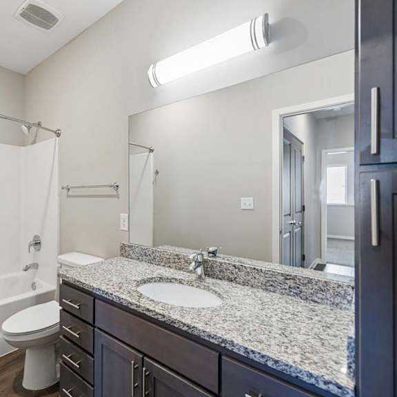 Thumbnail 19 of 36 Apartment bathroom with a granite-look single-sink vanity, dark cabinetry, a large mirror, and a tub-shower combination with a white surround.
