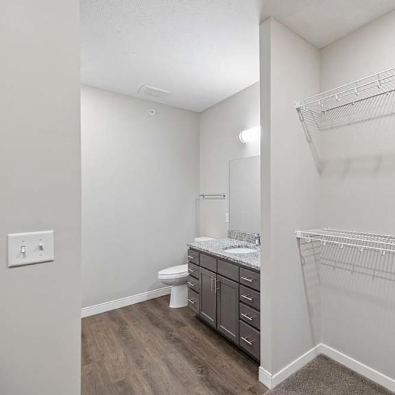 Thumbnail 32 of 36 Walk-in apartment closet with built-in wire shelving and an adjacent bathroom featuring a single-sink vanity, dark cabinetry, and wood-style flooring.