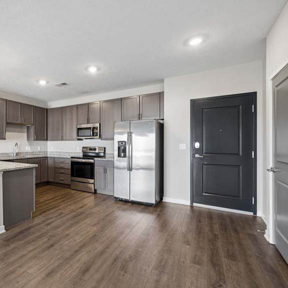 Thumbnail 23 of 36 Open-concept apartment kitchen with a large island, dark cabinetry, granite-look countertops, stainless steel appliances, pendant lighting, and wood-style flooring.