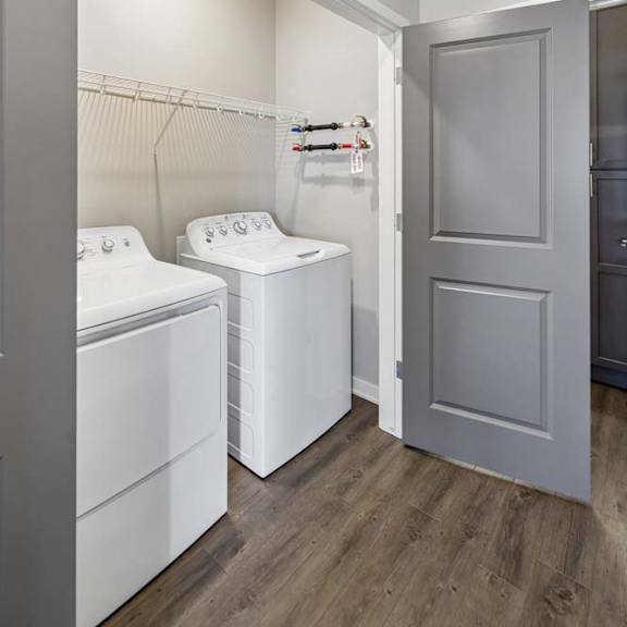 Thumbnail 35 of 36 Apartment laundry room with side-by-side washer and dryer, built-in wire shelving, wood-style flooring, and a gray interior door leading to the hallway.