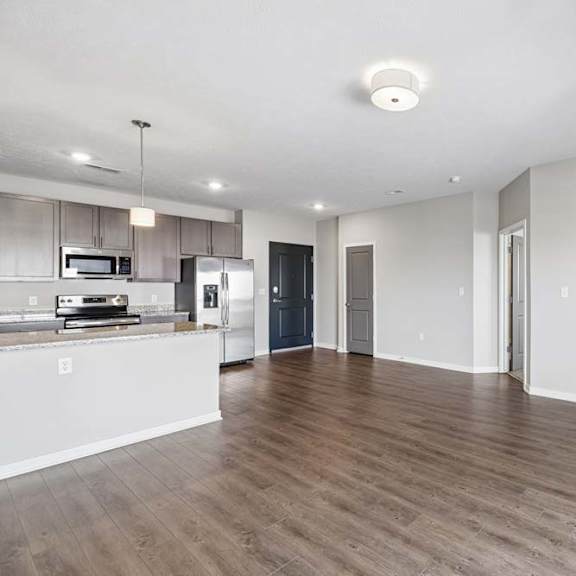Thumbnail 25 of 36 Spacious open-concept apartment living room with wood-style flooring, abundant natural light, and a modern kitchen featuring a breakfast bar, dark cabinetry, and stainless steel appliances.