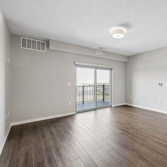 Thumbnail 26 of 36 Spacious open-concept apartment living room with wood-style flooring, a large sliding glass door leading to a private balcony, and an adjoining kitchen with a breakfast bar.
