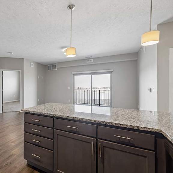 Thumbnail 22 of 36 Open-concept apartment living area viewed from the kitchen island, featuring wood-style flooring, pendant lighting, and a large sliding glass door leading to a private balcony.