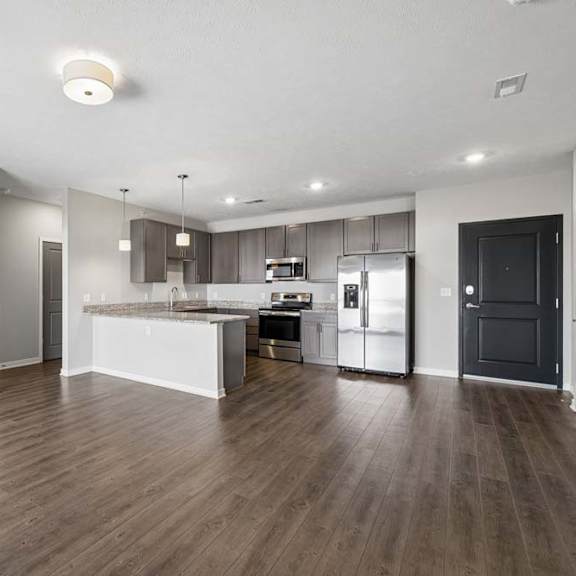 Thumbnail 24 of 36 Spacious open-concept apartment living area with wood-style flooring, a modern kitchen featuring a large island, dark cabinetry, stainless steel appliances, and recessed lighting.