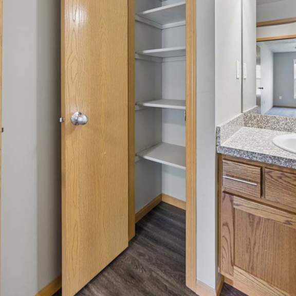 Thumbnail 11 of 13 Built-in apartment pantry with open shelving, located beside the bathroom vanity with wood cabinetry and a stone-look countertop.