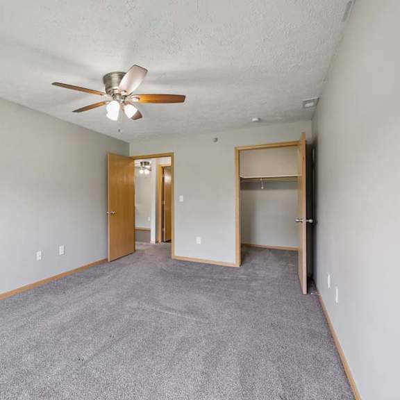 Thumbnail 7 of 13 Spacious apartment bedroom with soft carpeting, a ceiling fan, an open closet, and access to an adjoining hallway.