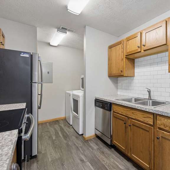 Thumbnail 3 of 13 Galley-style apartment kitchen with wood cabinetry, a subway tile backsplash, stainless steel appliances, and an adjacent in-home laundry room.