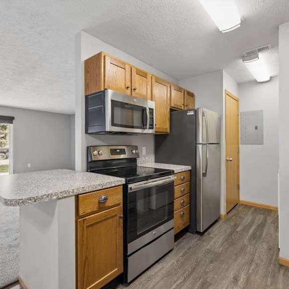 Thumbnail 2 of 13 Apartment kitchen with wood cabinetry, stainless steel appliances, a breakfast bar, and an open layout overlooking the living room.