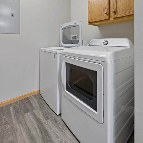 Thumbnail 12 of 13 Dedicated apartment laundry room with a side-by-side washer and dryer, overhead storage cabinets, and wood-style flooring.