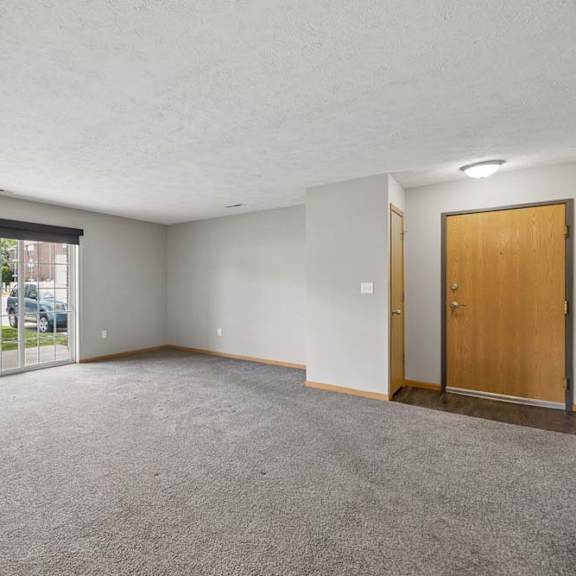 Thumbnail 6 of 13 Spacious apartment living room with carpeted flooring, a large sliding glass door, and an open entryway with natural light.