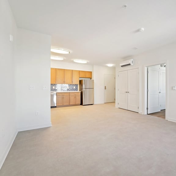 a spacious, empty room with white walls and a kitchen area in the background.