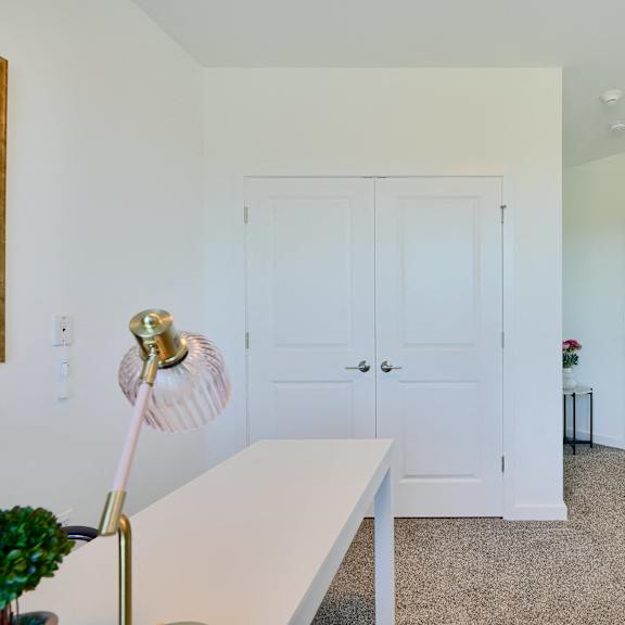 now leasing senior 1-bedrooms in baltimore, md. a white table with a gold lamp stands in a room with white walls and a carpeted floor.