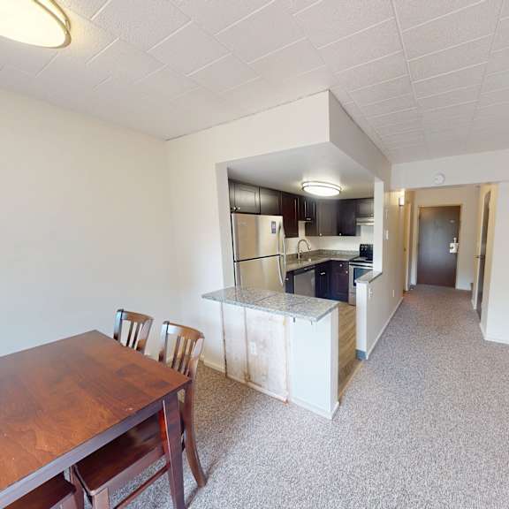 a room with a wooden table and chairs in the foreground and a kitchen area in the background.