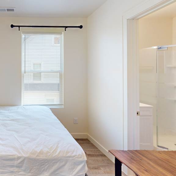 a bedroom with a bed, a window with blinds, a wooden table, and a glass shower stall.