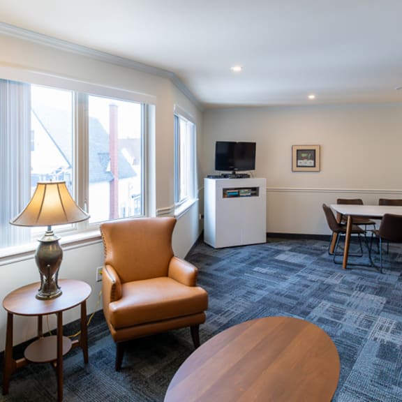 a room with a carpeted floor, a sofa, a chair, a table, a lamp, a television, and a fridge.