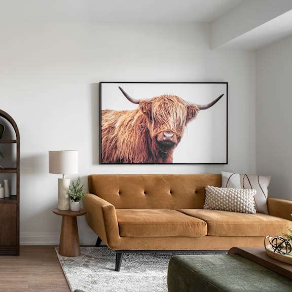a living room with a tan couch, a brown side table, a lamp, a plant, a framed picture of a highland cow, and a wooden cabinet.