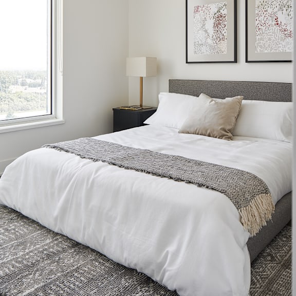 a bed with a white comforter and a grey blanket is in the foreground with a lamp and two pictures on the wall.