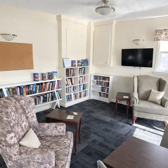 a living room with a grey floral armchair, a wooden coffee table, a grey sofa, a bookshelf with books, a television, and a window with curtains.