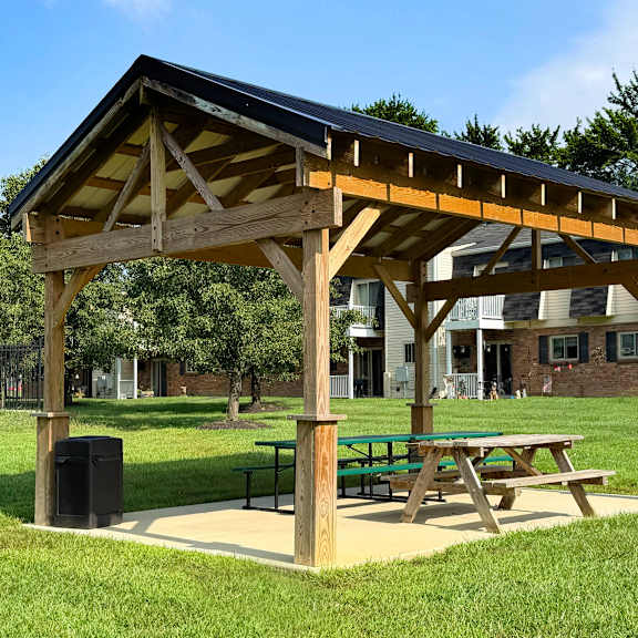 a wooden pavilion with a black roof is situated in a grassy area with a picnic table and a trash can.