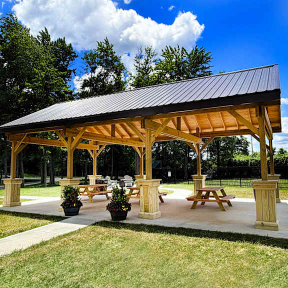 a wooden pavilion with a black roof and four pillars is surrounded by grass and trees.
