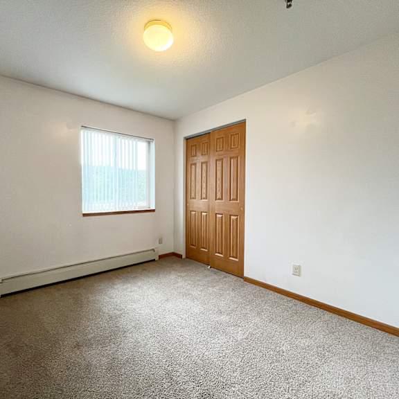 a room with a brown carpet, a window with white blinds, a wooden door, and a white wall.