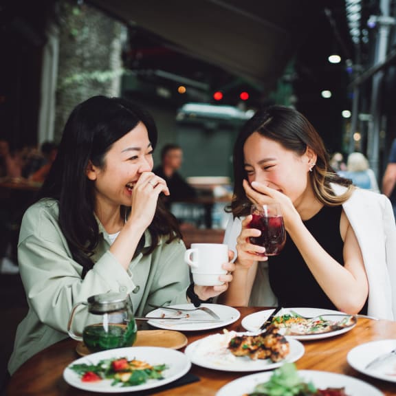 Two ladies laughing and enjoying lunch at an al fresco spot, holding their drinks.