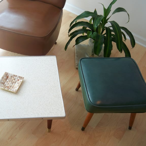 A brown leather chair sits next to a white table with a cracker on it and a green chair with a plant in the background.