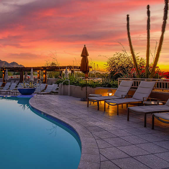 a poolside area with lounge chairs and a mountain in the background during sunset.
