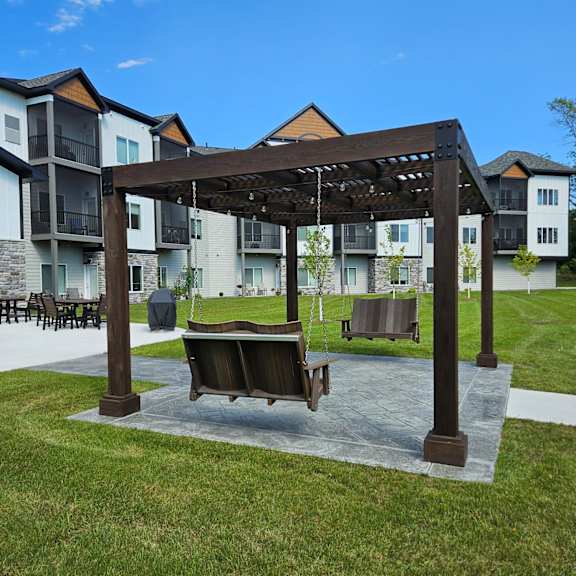 a wooden pergola with a hanging bench is in the foreground of a grassy area with apartment buildings in the background.