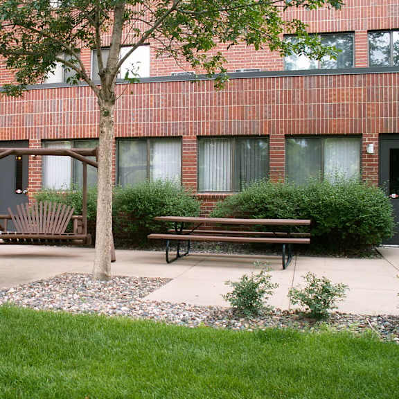 a tree stands in front of a brick building with a swing set and two benches.