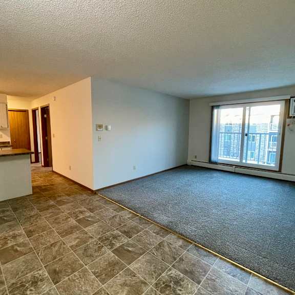 a spacious living room with a kitchen area to the left and a window with a view of a building outside.