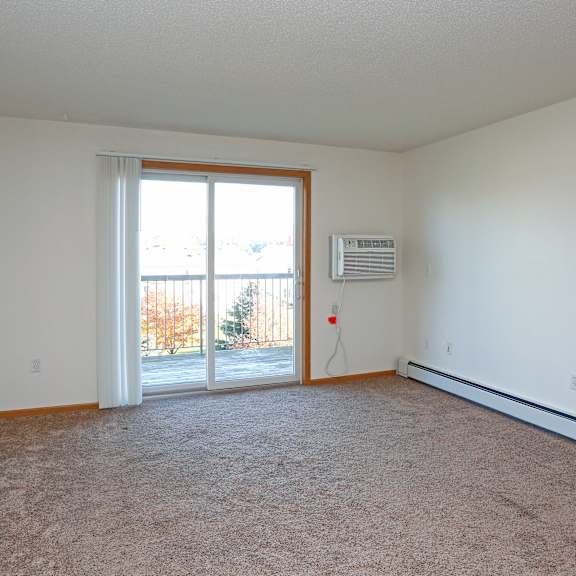 a room with a carpet floor, a window, a door, and a wall-mounted air conditioner unit.