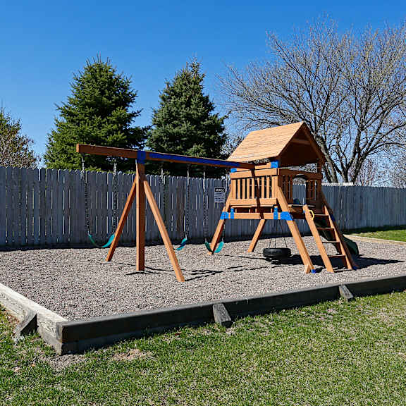 a wooden swing set with a brown roof is situated on a gravel area in a backyard.