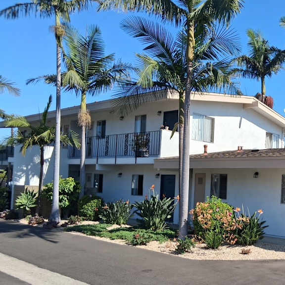 a white two-story house with a blue door and windows surrounded by palm trees.