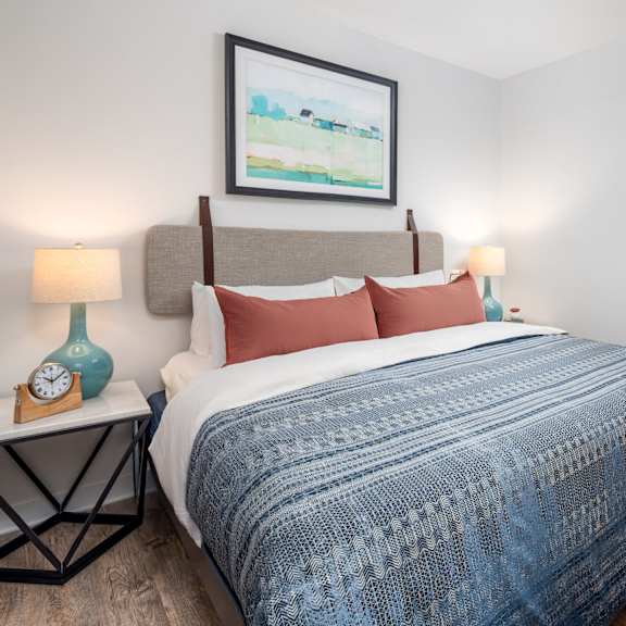 a bed with a blue blanket and red pillows is in the foreground of a room with a painting on the wall and a nightstand with a clock and a vase.
