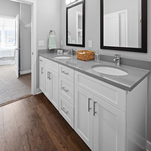 a bathroom with a double vanity, two mirrors, ample cabinet space, and hardwood-style flooring near a doorway to a walk-in closet and bedroom.