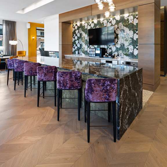 a community kitchen with a long bar, modern purple barstools, pendant chandelier, and a tv in a resident lounge.