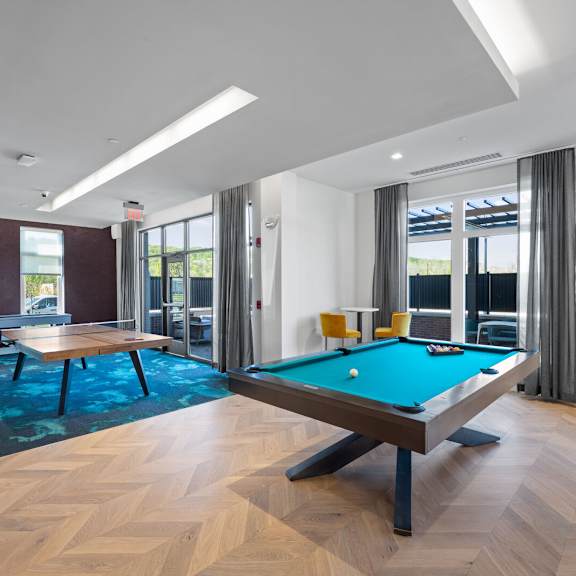 a clubhouse with a billiards table, a ping pong table, and a shuffleboard near doors leading to an outdoor community lounge.